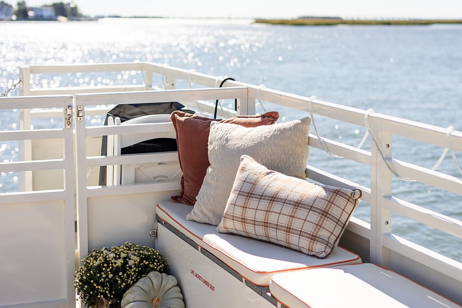 A plaid pillow, mums and a white pumpkin on the deck bench by the rail